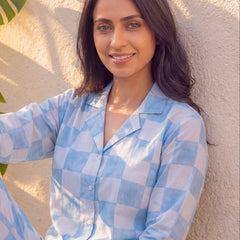 Woman wearing a blue checkered outfit sitting against a textured wall with leaves in the background