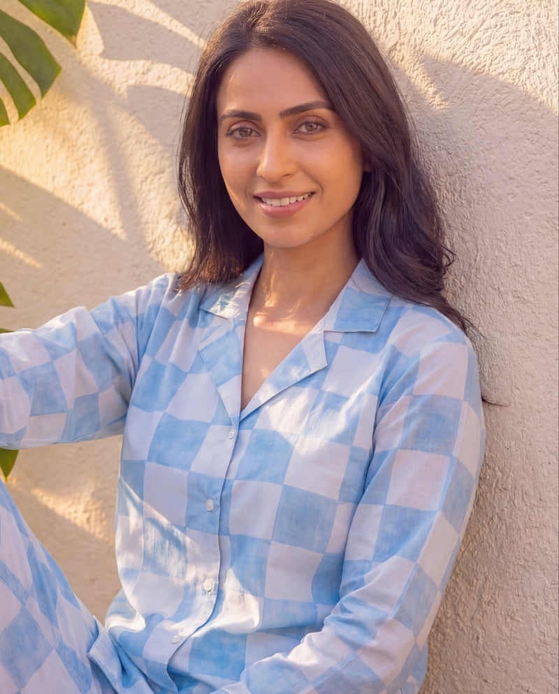Woman wearing a blue checkered outfit sitting against a textured wall with leaves in the background